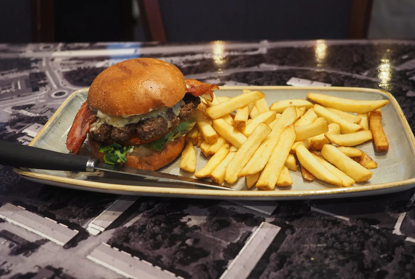Burger mit Rindfleisch, Käse und Bacon auf einem länglichen Teller, daneben Pommes frites