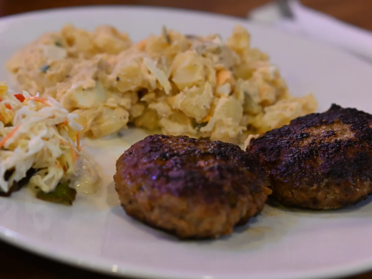 Zwei gebratene Hacksteaks mit Kartoffelsalat und Krautsalat auf einem weißen Teller