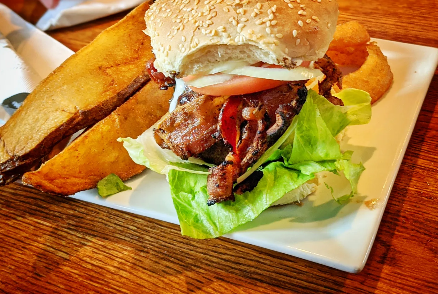 Burger mit Bacon, Tomate und Salat im Sesambrötchen, daneben Kartoffelspalten auf einem weißen Teller