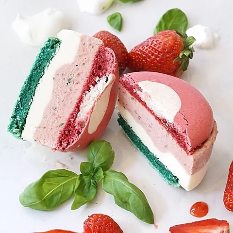 Zwei aufgeschnittene Macaron-Eis-Sandwiches mit Erdbeer- und Vanilleeis zwischen bunten Baiserhälften, daneben frische E