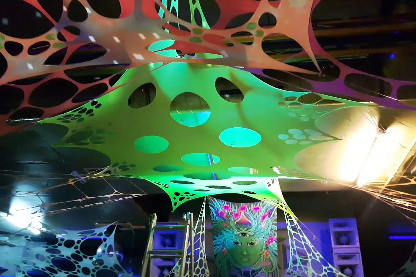 Fluoreszierende Spanntücher in Grün und Orange unter der Decke des Juice Club, dahinter Lautsprecher und ein bemaltes Banner