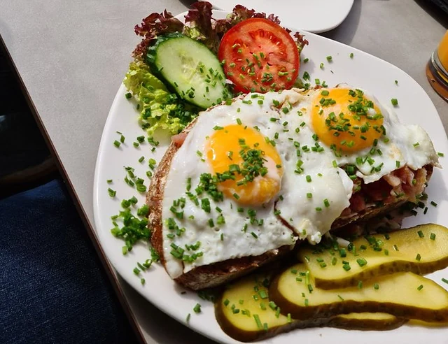 Strammer Max mit zwei Spiegeleiern, Schnittlauch, Gurke und Tomate, dahinter ein Erdbeertörtchen