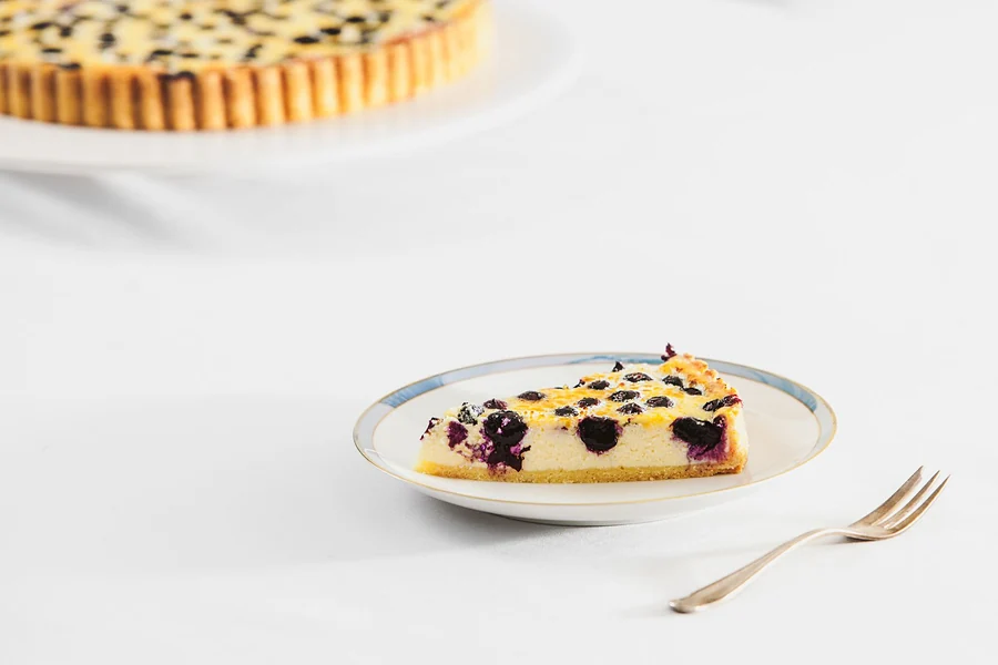 Stück Heidelbeer-Käse-Tarte auf kleinem Teller, dahinter die ganze Tarte