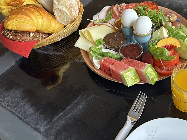 Frühstücksplatte mit gekochten Eiern, Käse, Aufschnitt, Wassermelone, Tomaten und Aufstrichen, dahinter ein Korb mit Croissant und Brötchen