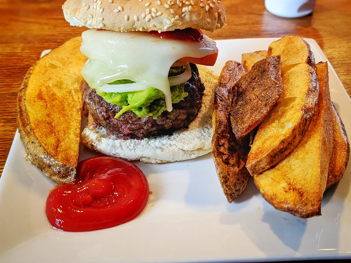Burger mit Rindfleisch, geschmolzenem Käse und Guacamole, daneben Kartoffelspalten und Ketchup