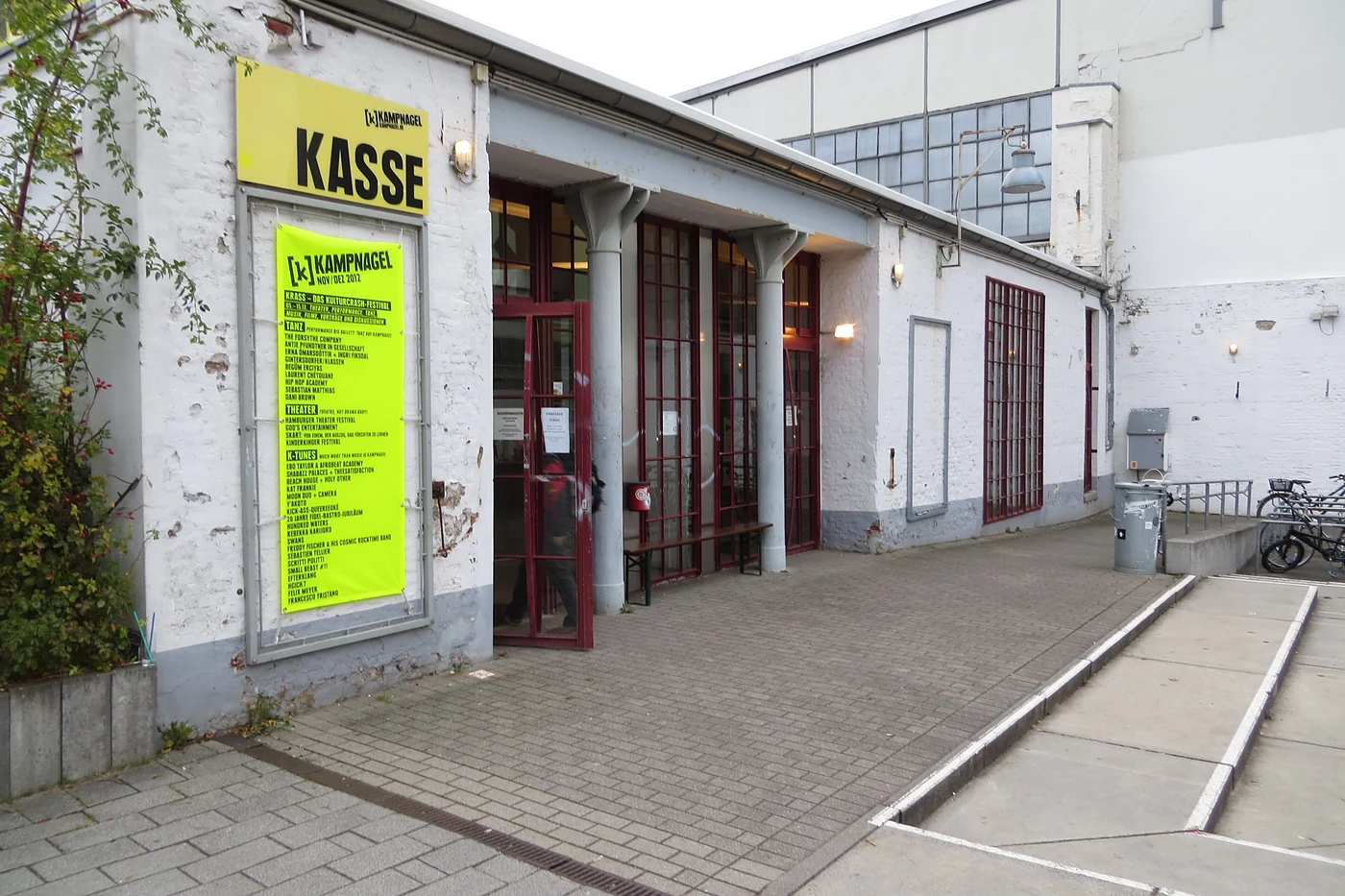 Eingang zur Kasse von Kampnagel mit gelbem Plakat und rotem Tor in der weiß gestrichenen Halle