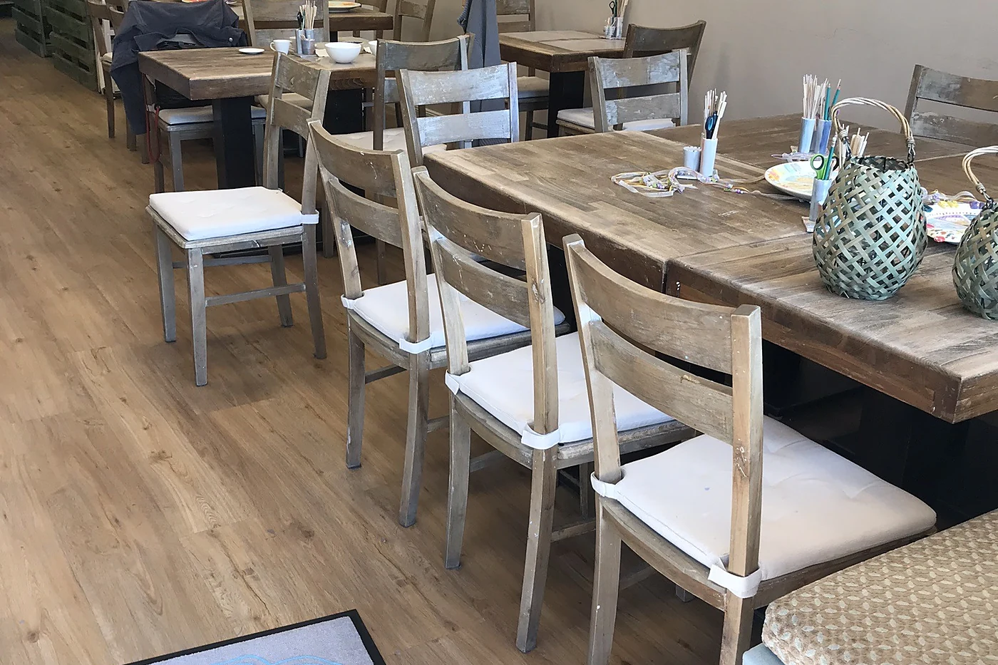 Heller Raum mit Holztischen, weißen Stühlen, Farbtöpfen auf dem Tisch und Bildern an der Wand