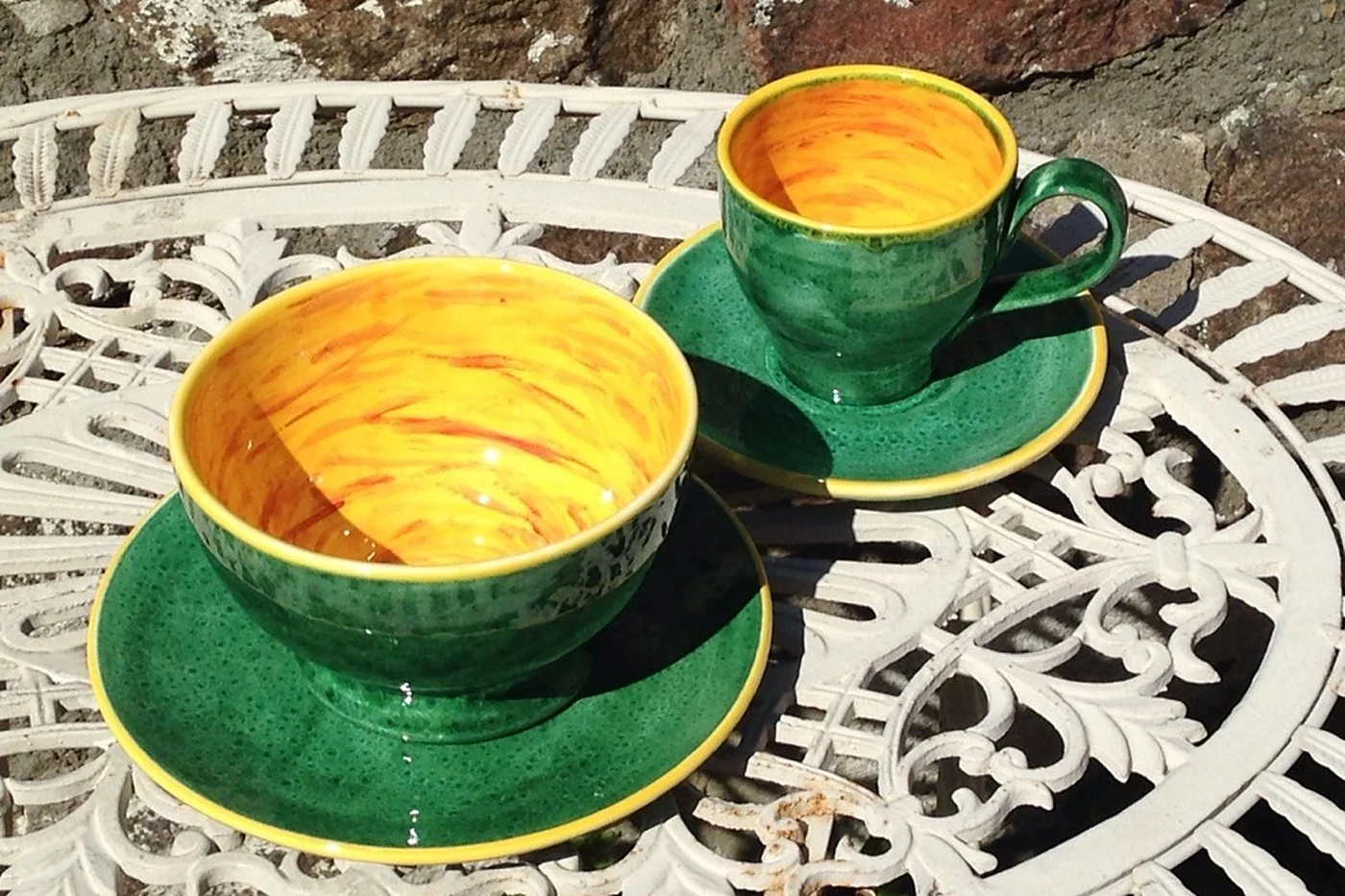 Selbst bemalte Keramik nach dem Brennen: gelb-grüne Schale und Tasse mit Untertasse auf einem weißen Gartentisch