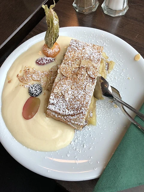 Blätterteigschnitte mit Puderzucker auf Vanillesauce mit Physalis, Weintraube und Blaubeere auf einem weißen Teller