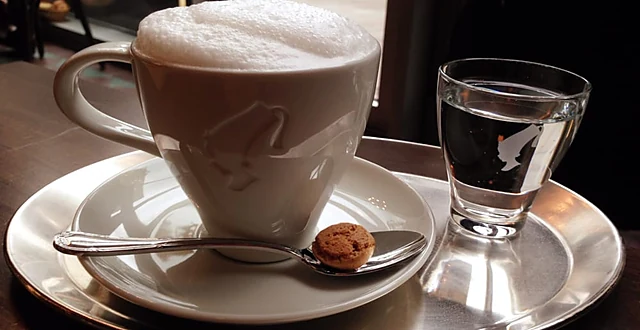 Cappuccino in weißer Tasse mit Milchschaum, dazu ein Glas Wasser und ein Keks auf einem silbernen Tablett