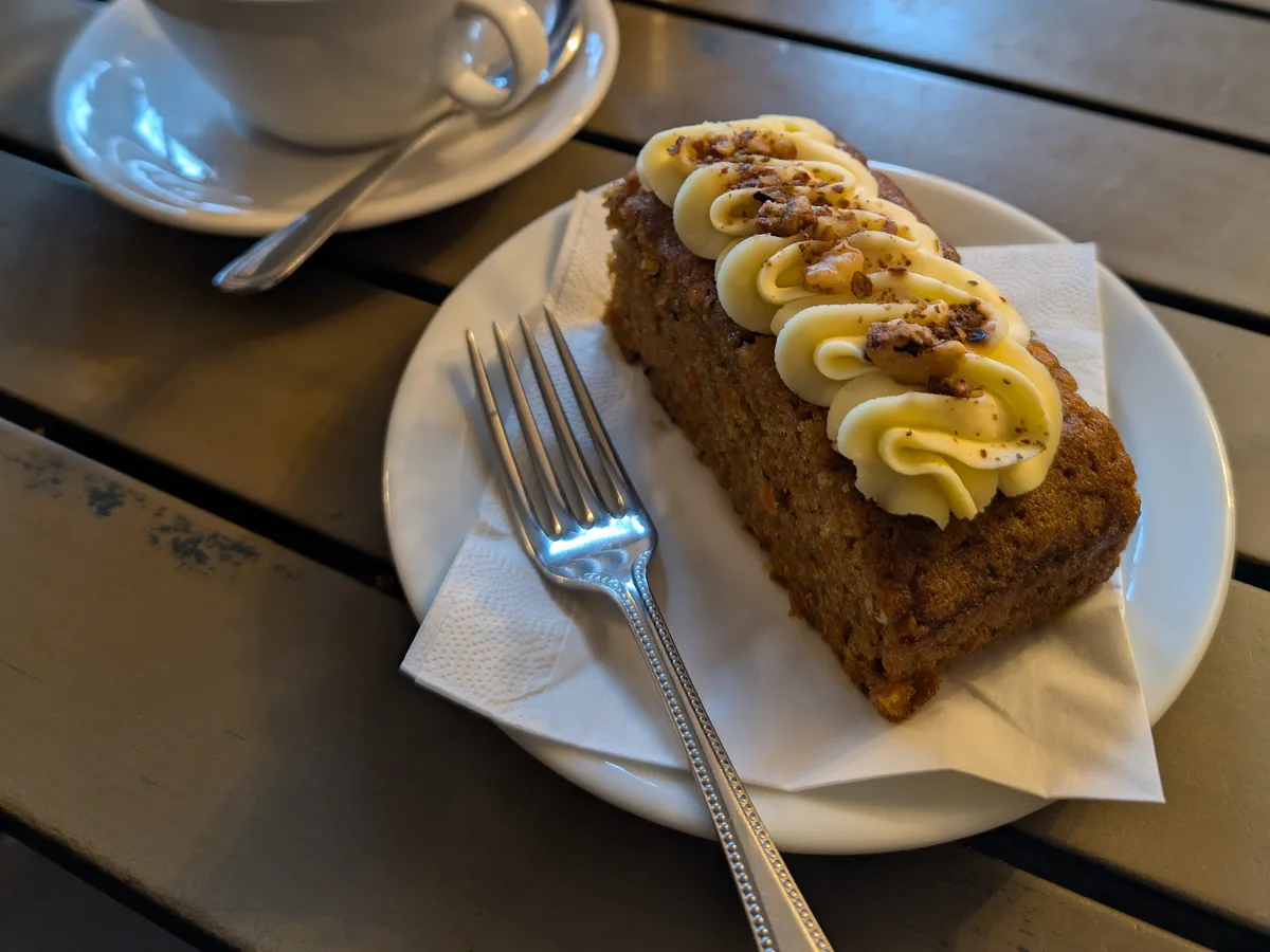 Stück Möhrenkuchen mit Frischkäsecreme auf einem Teller, daneben ein Milchkaffee