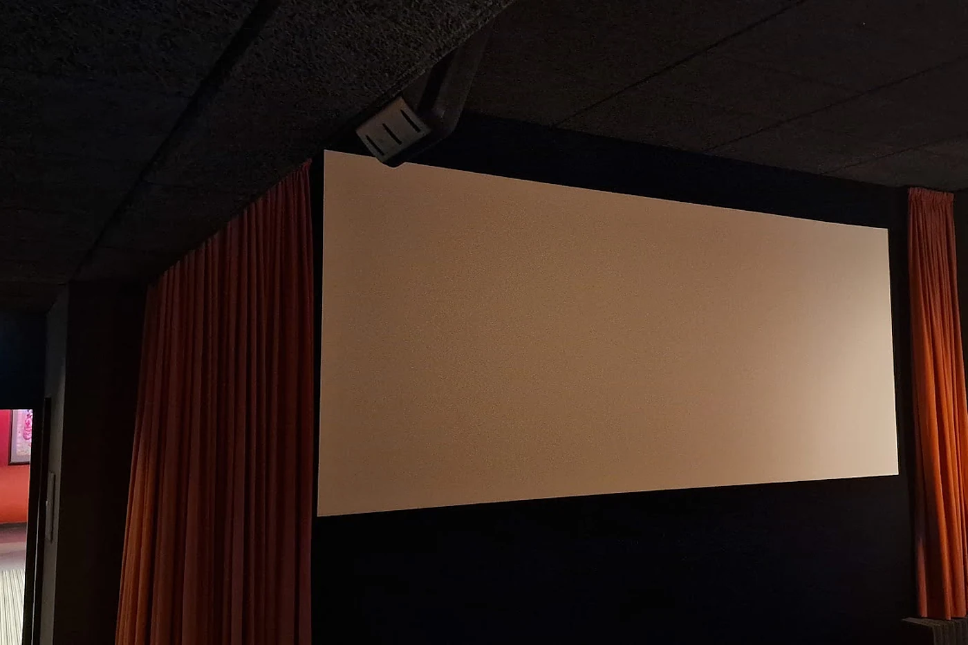 Kleinerer Kinosaal mit heller Leinwand zwischen orangefarbenen Vorhängen und dunkler Wandverkleidung