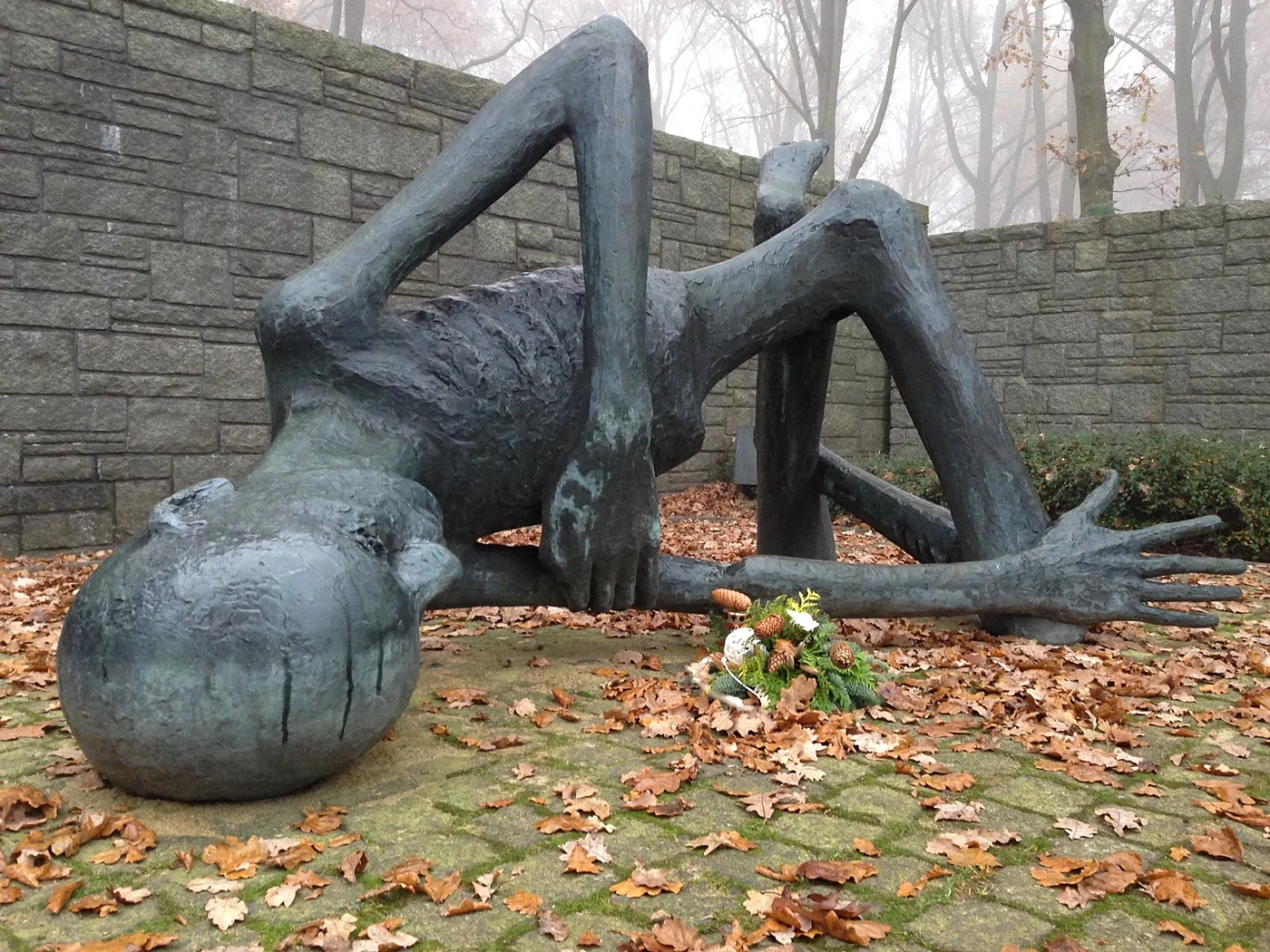 Bronzeskulptur eines ausgemergelten, am Boden liegenden Häftlings vor einer Natursteinmauer, davor ein kleiner Blumenstr
