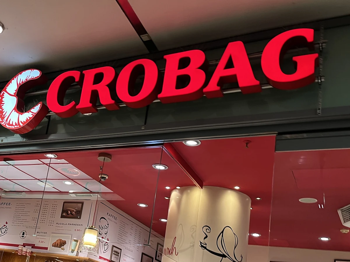 Roter Leuchtschriftzug CROBAG mit stilisiertem Croissant über dem Eingang der Filiale