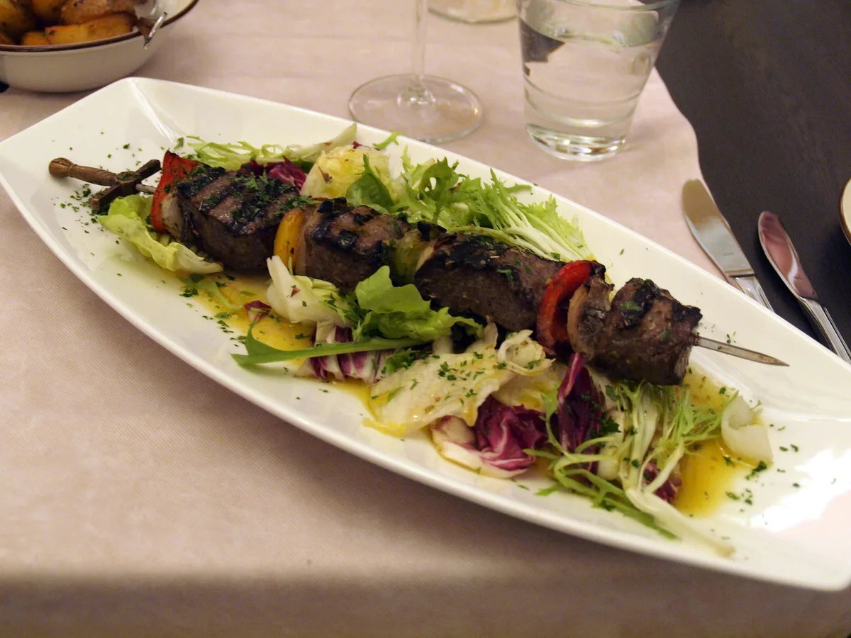 Gegrillter Fleischspieß mit Paprika und Zwiebeln auf einem länglichen weißen Teller mit Blattsalaten