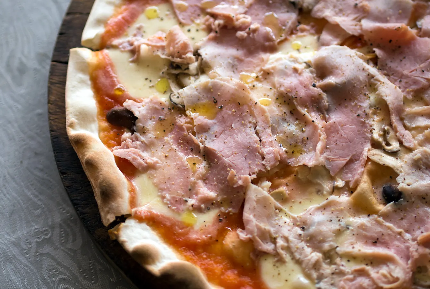 Dünn ausgerollte Pizza mit Kochschinken, Mozzarella und Pilzen auf einem Holzbrett