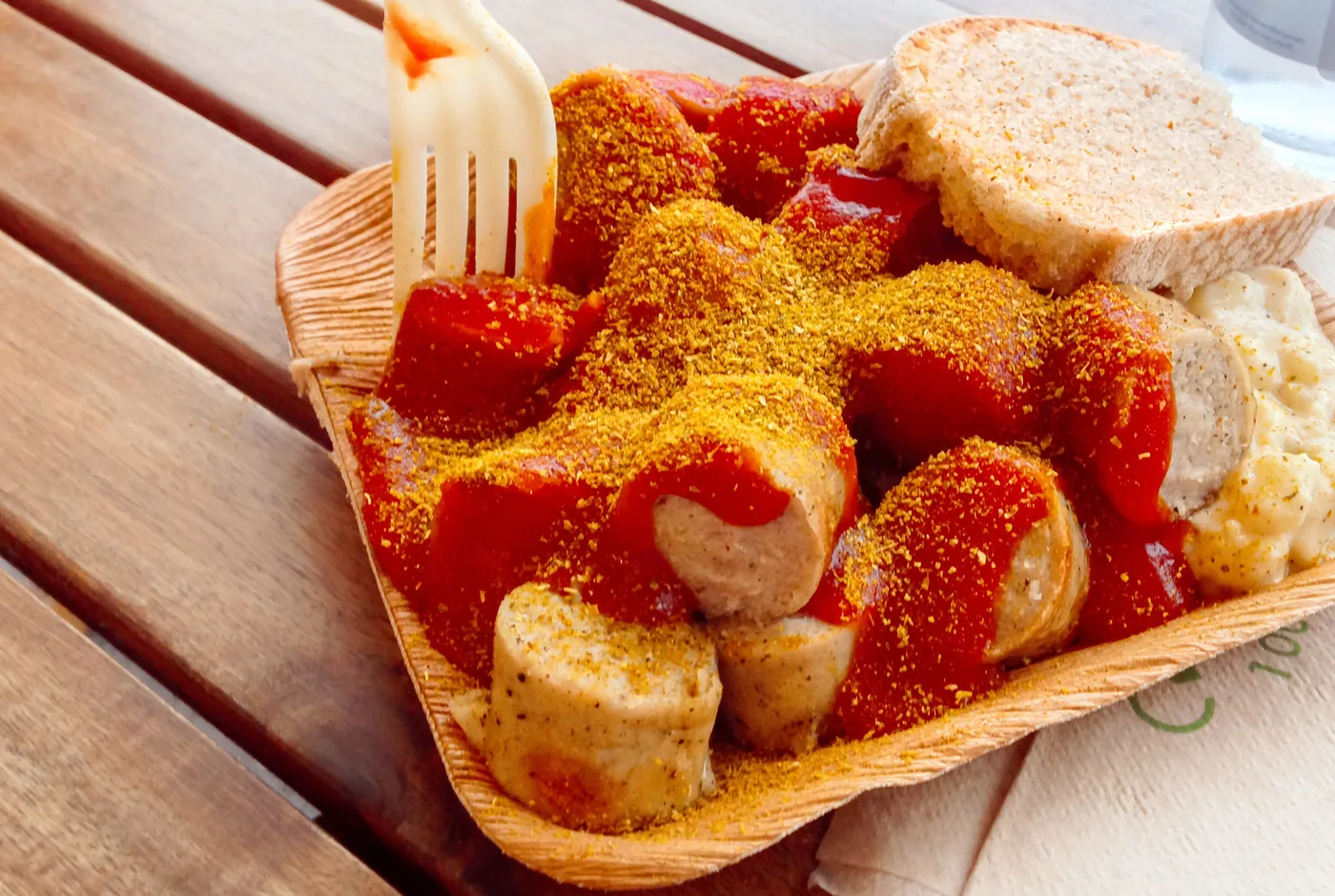 Currywurst mit Tomatensauce und Currypulver in einer Pappschale, dazu ein Stück Brot