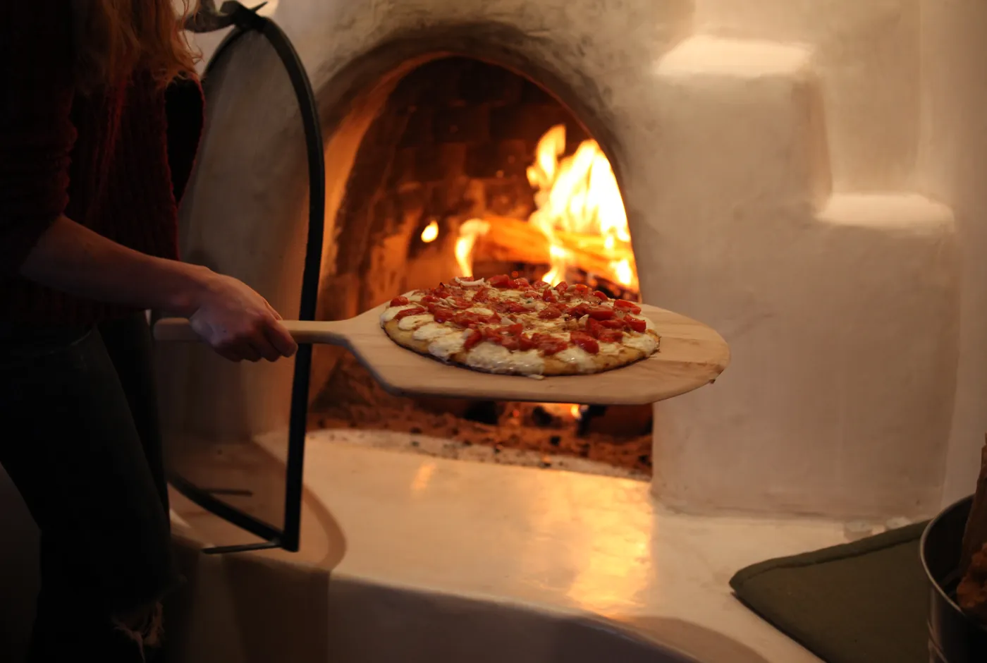 Belegte Pizza auf einem Holzschieber vor einem brennenden Holzofen