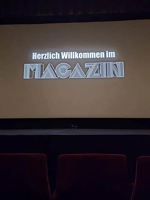 Kinoleinwand mit dem projizierten Schriftzug Herzlich Willkommen im Magazin, davor rote Sessellehnen