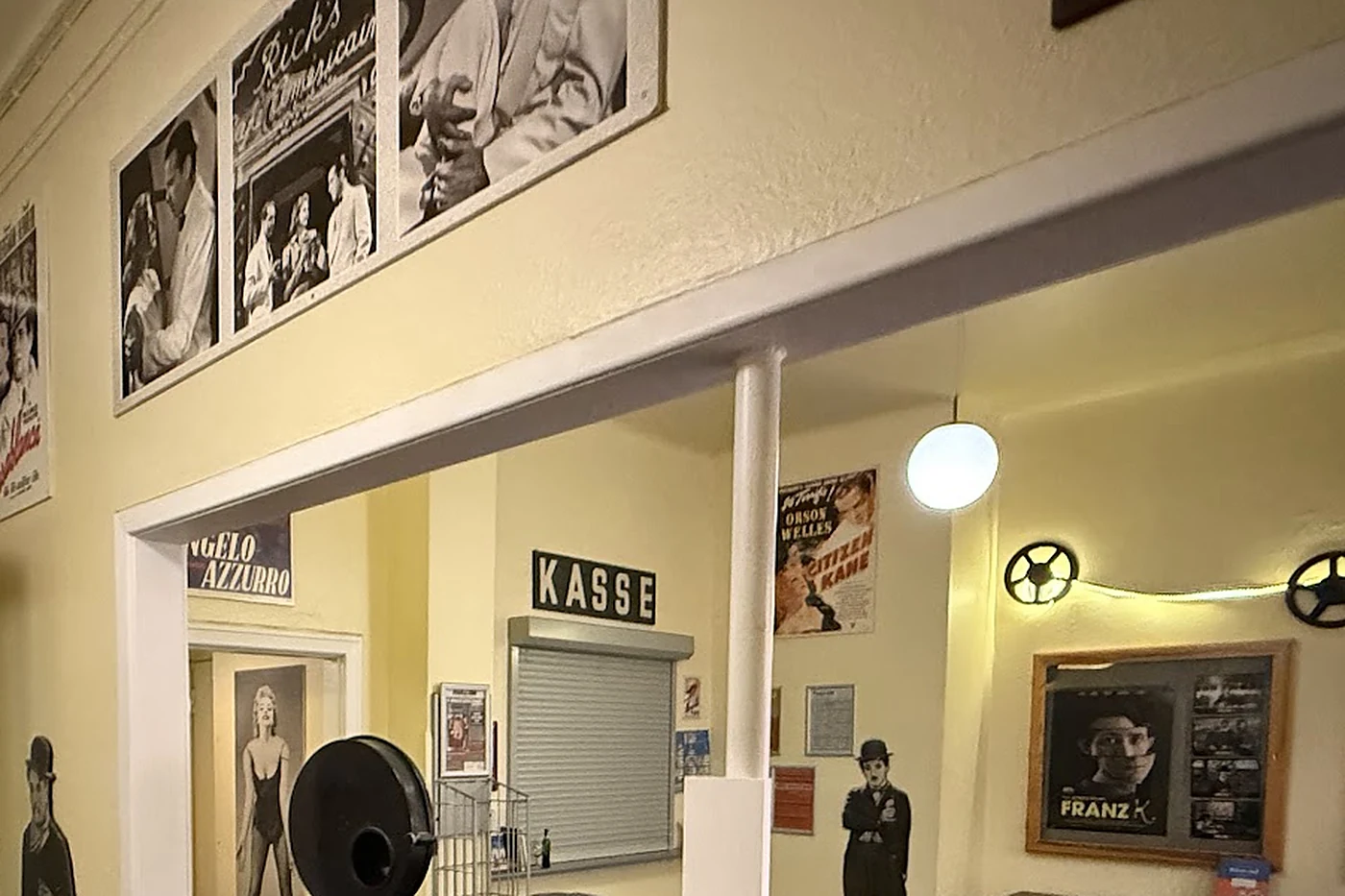 Kinofoyer mit Schild Kasse über einem Rollladenfenster, altem Filmprojektor und gerahmten Filmfotos an gelber Wand