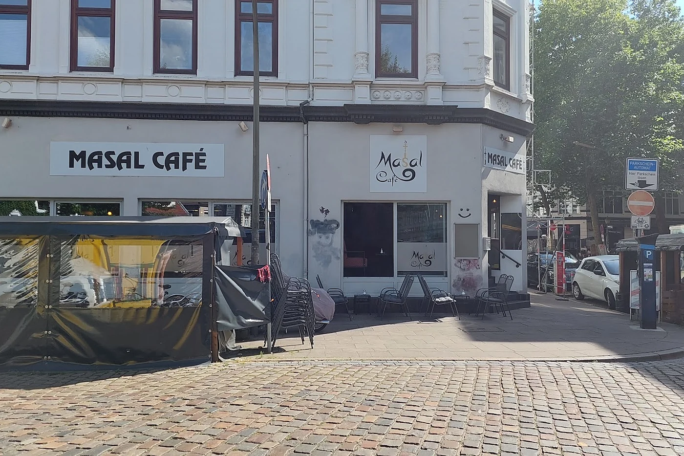 Weißes Eckhaus mit dem Schild Masal Café, davor Kopfsteinpflaster, Stühle und ein überdachter Außenbereich
