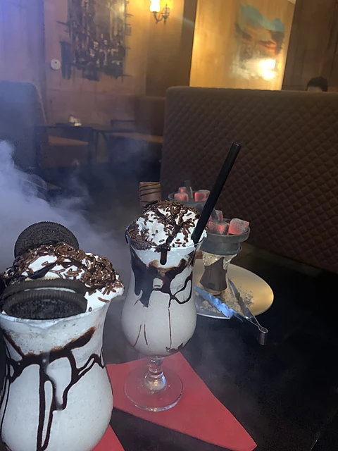 Zwei Milchshakes mit Sahnehaube, Schokoladensauce und Keksen auf einem dunklen Tisch, daneben ein Shisha-Kopf, im Hintergrund Rauchschwaden