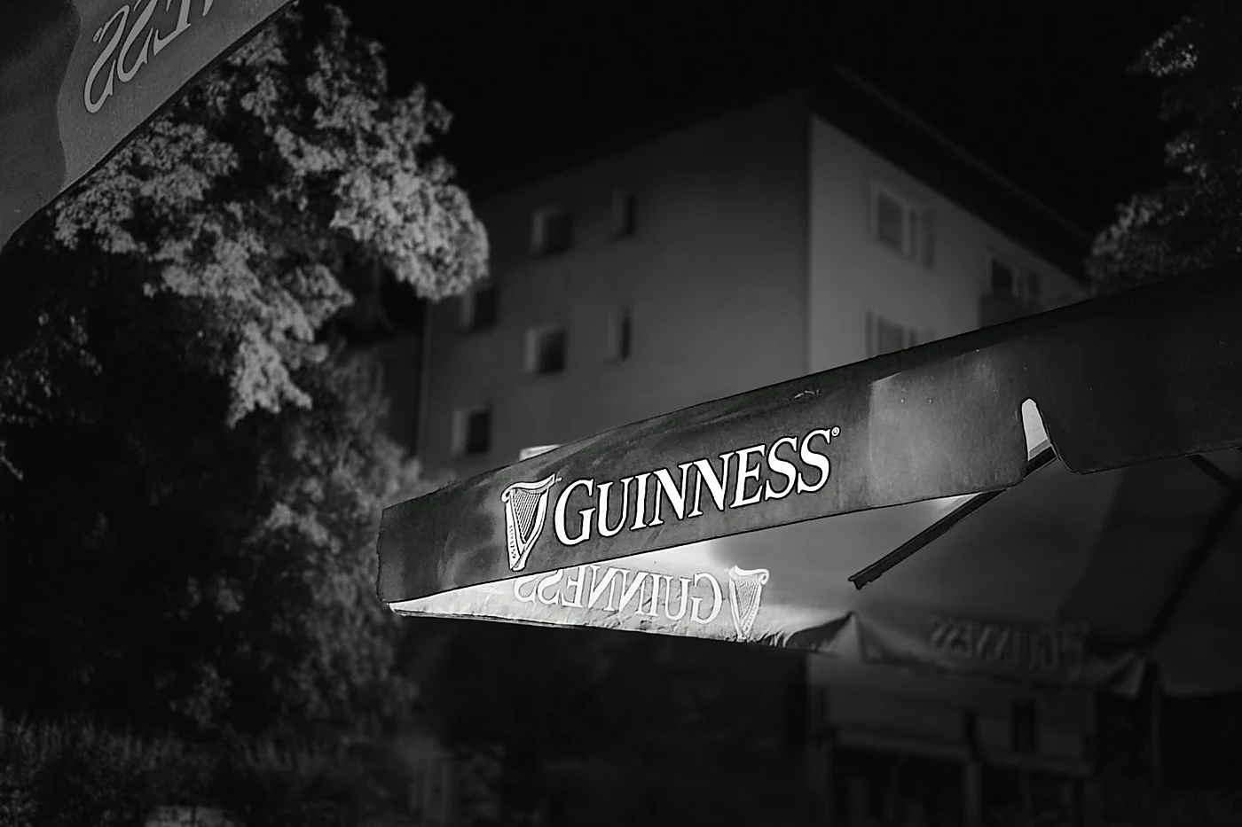 Schwarz-Weiß-Aufnahme eines Guinness-Sonnenschirms bei Nacht vor Bäumen und einer Hausfassade