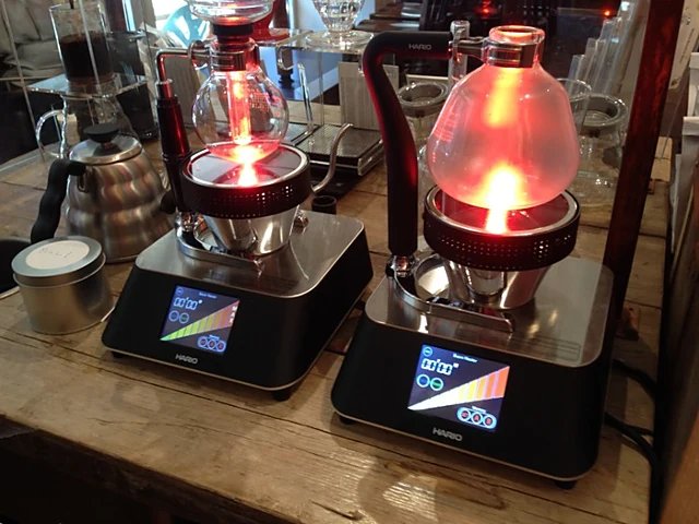 Zwei Syphon-Kaffeebereiter mit rot leuchtenden Halogenbrennern auf digitalen Waagen an der Brew-Bar