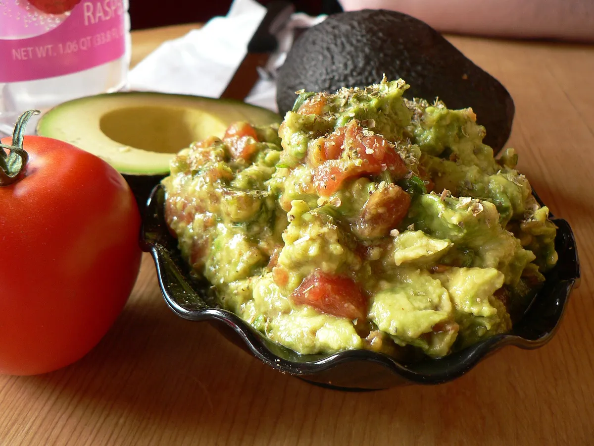 Guacamole in einem Steinmörser, daneben eine halbierte Avocado und eine Tomate