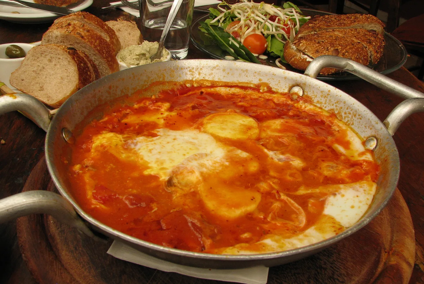 Shakshuka in einer Pfanne mit Tomatensauce und Eiern, daneben Brot und Salat