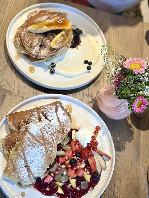 Zwei Teller mit Pancakes, Beerensauce, Creme und frischen Früchten auf einem Holztisch, daneben eine kleine Blumenvase