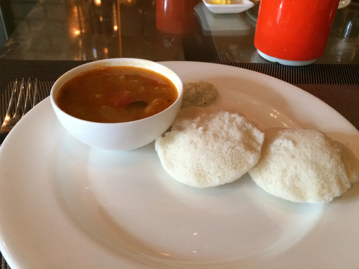 Zwei Idli auf einem weißen Teller, daneben eine Schale Sambar und Kokoschutney