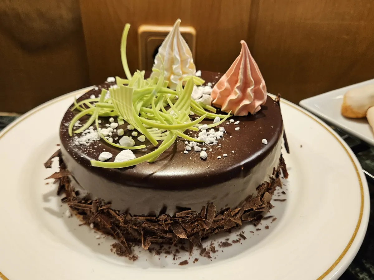 Schokoladentorte mit Baiserspitzen auf einem weißen Teller, im Hintergrund weiteres Gebäck