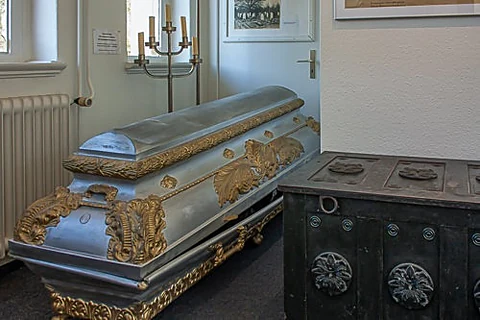 Historischer silberner Prunksarg mit vergoldeten Beschlägen neben einer schwarzen Truhe in der Ausstellung