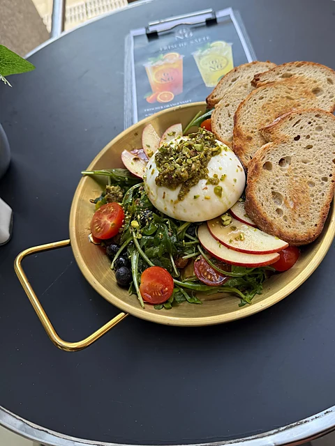 Goldene Schale mit Burrata, Pesto, Rucola, Kirschtomaten, Apfelscheiben und Oliven, dazu geröstetes Brot auf einem schwarzen Tisch draußen