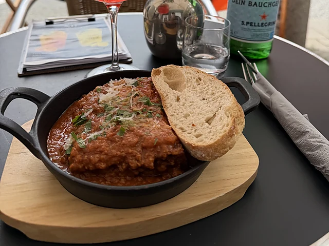 Gusseiserne Pfanne mit Hackfleisch-Ragout und Kräutern, dazu ein Stück Brot auf einem Holzbrett, im Hintergrund ein Aperol Spritz