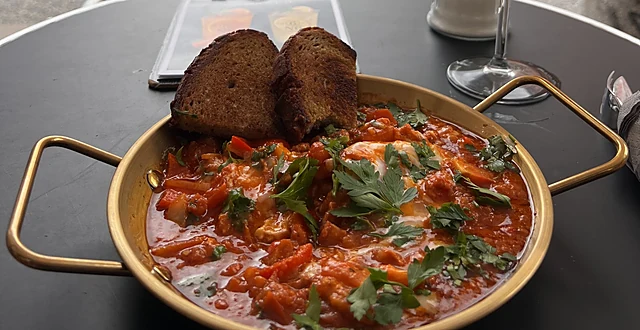 Goldene Pfanne mit Tomaten-Schakschuka und Petersilie, darauf zwei Scheiben dunkles Brot