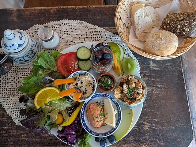 Frühstücksteller mit Salat, Dips, Tomaten, Gurke, Orange und Brötchenkorb auf einem Holztisch mit Häkeldecke