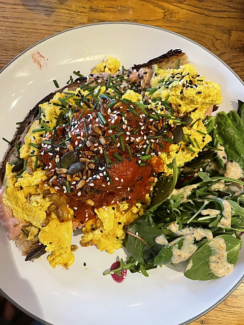 Geröstetes Sauerteigbrot mit Rührei, Tomatensauce, Sesam, Kürbiskernen und Schnittlauch, daneben Rucola auf weißem Telle