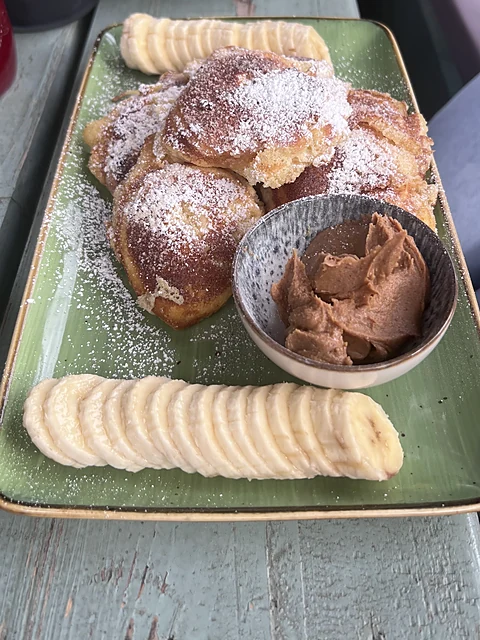 Kleine Pancakes mit Puderzucker auf grünem Teller, dazu Bananenscheiben und ein Schälchen Schokocreme