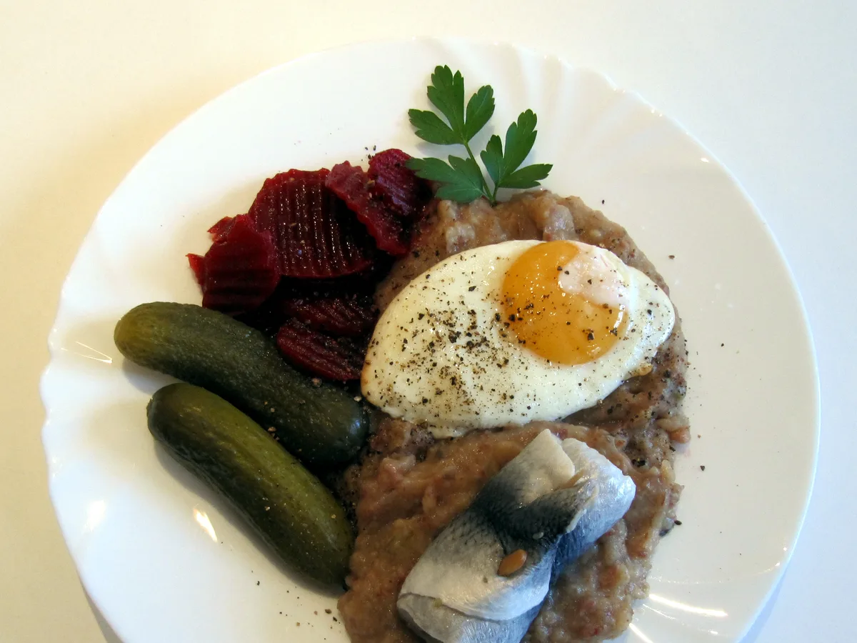 Labskaus mit Spiegelei, Rollmops, Roter Bete und Gewürzgurken auf einem weißen Teller
