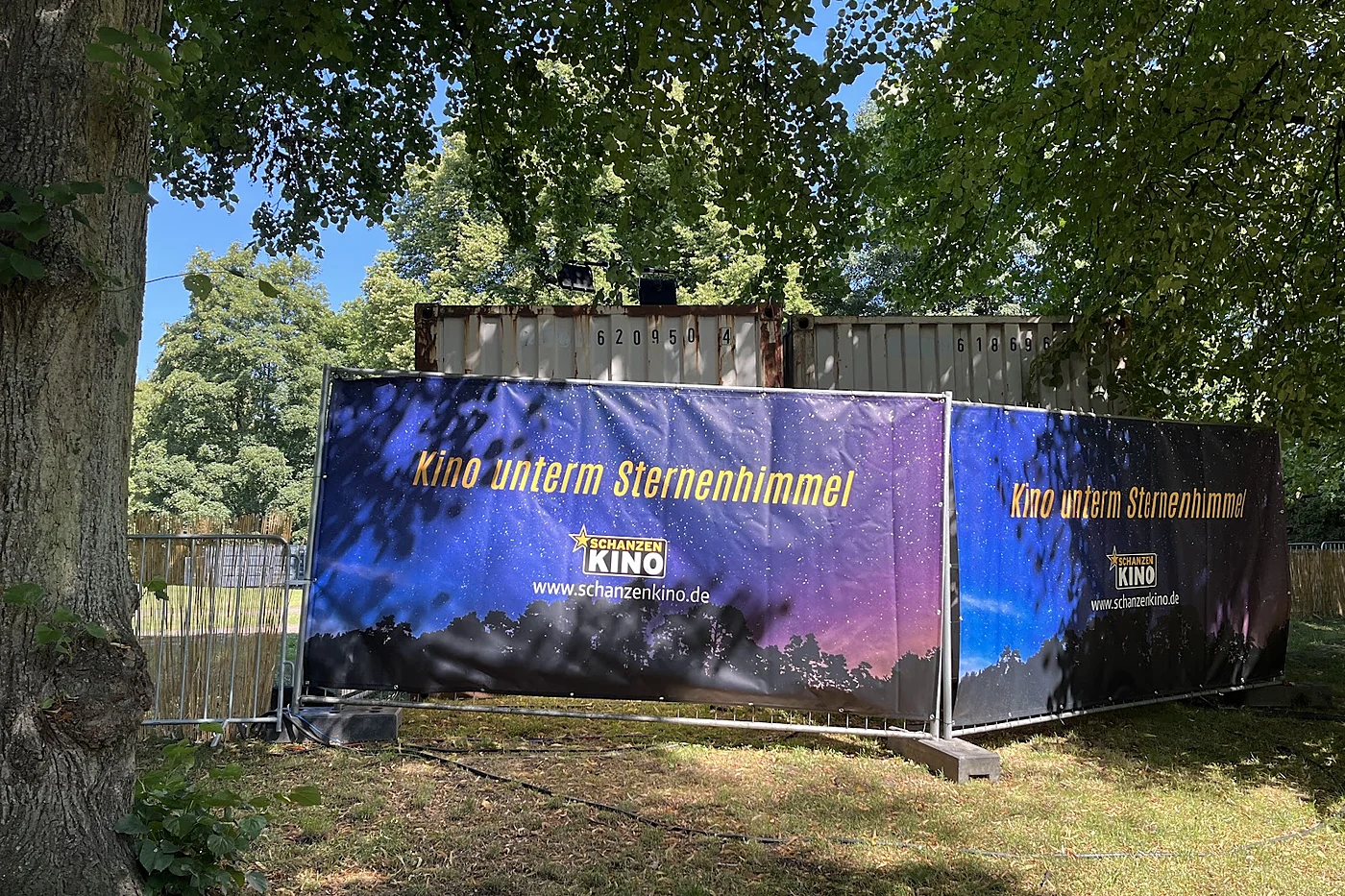Bauzaunbanner mit Sternenhimmelmotiv und der Aufschrift Kino unterm Sternenhimmel vor Überseecontainern im Park
