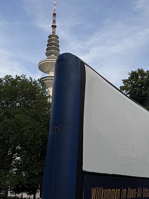 Seitenansicht einer aufblasbaren Kinoleinwand im Park, dahinter der Heinrich-Hertz-Turm vor blauem Himmel