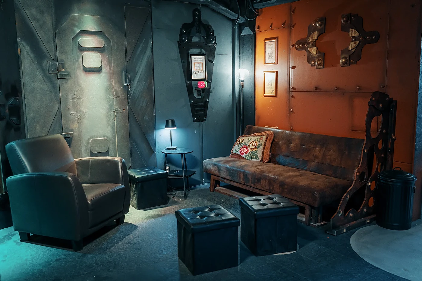 Lounge im Steampunk-Stil mit Ledersessel, Sofa, Hockern und einer Wand aus genieteten Metallplatten in blauem Licht