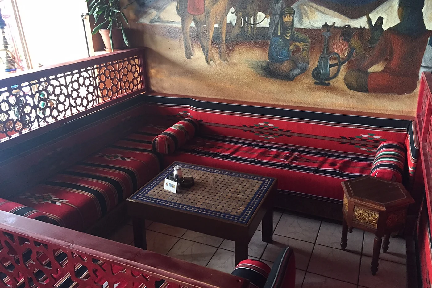 Orientalische Sitzecke mit roten Polstern, Holztisch mit Mosaikplatte und einem Wandgemälde mit Kamelen und Karawane