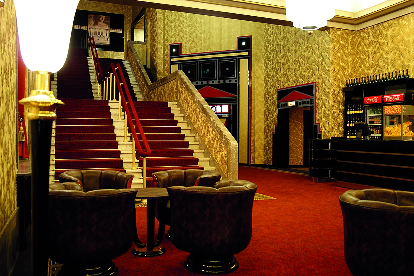 Kinofoyer mit goldener Tapete, roter Treppe mit Messinggeländer, Ledersesseln und Snacktheke