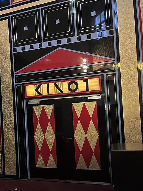 Schwarz-rot-goldene Art-déco-Tür mit Rautenmuster und beleuchtetem Schild mit der Aufschrift Kino 1