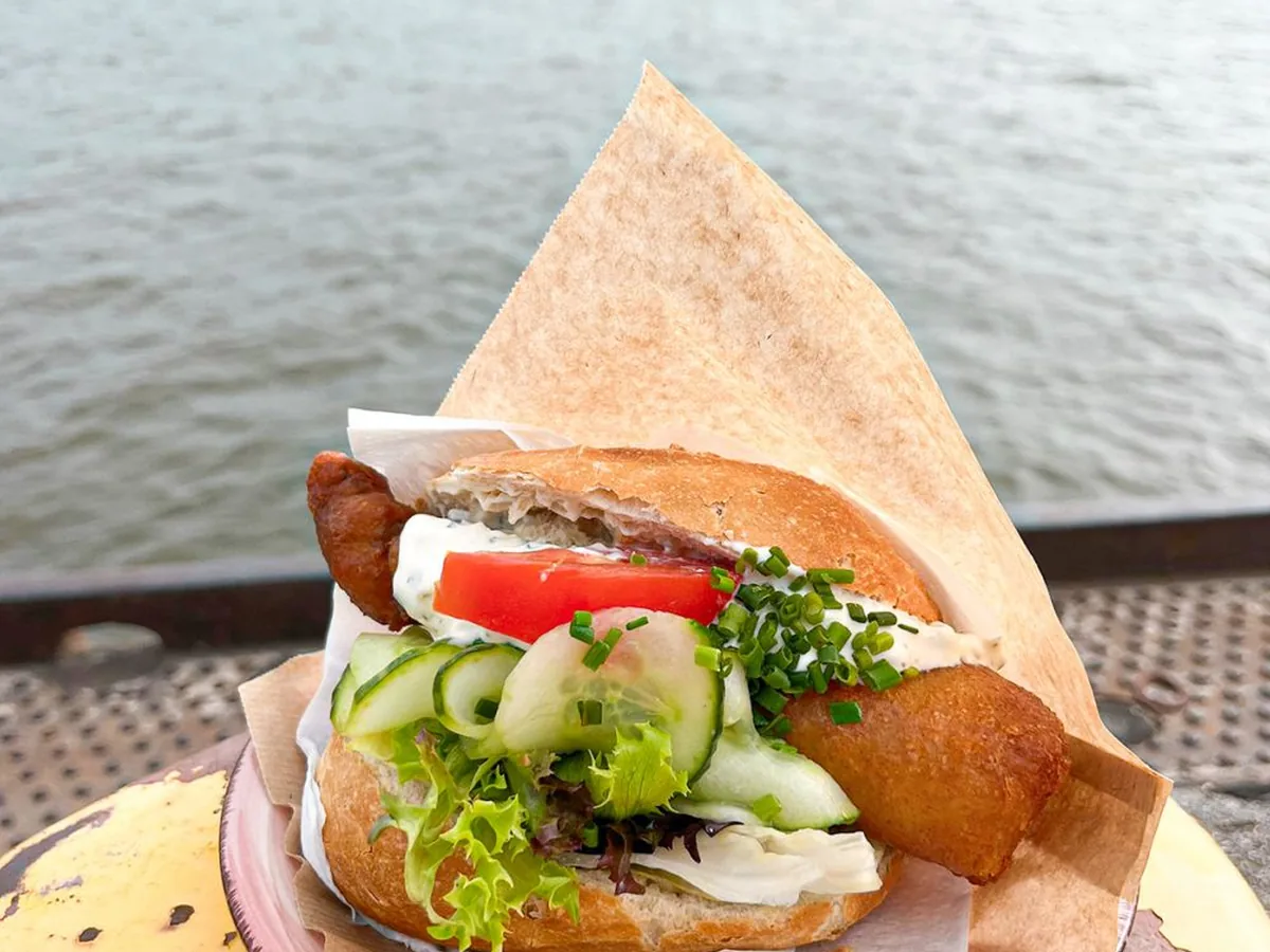 Fischbrötchen mit Backfisch, Gurke und Tomate auf einem gelben Poller an der Elbe