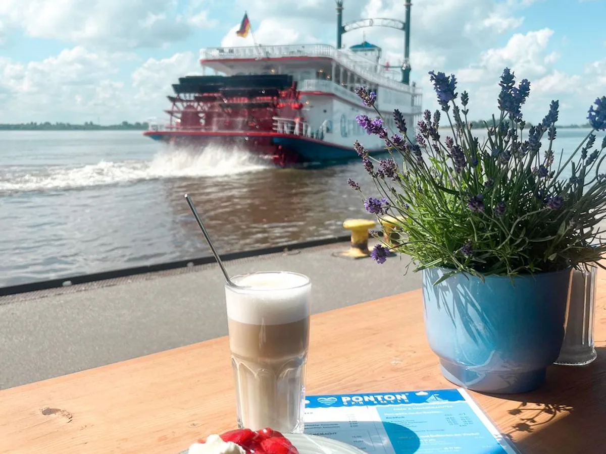 Latte Macchiato und Kuchen auf einem Tisch, dahinter ein Raddampfer auf der Elbe