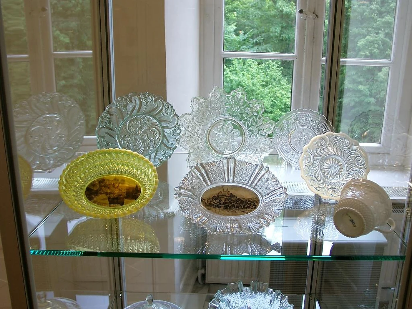 Glasvitrine mit gepressten Schalen und Tellern aus klarem, gelbem und opalweißem Glas mit Reliefmustern vor einem Fenste